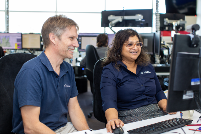 Two colleagues sit at desks, looking at a computer monitor and smiling in an office with multiple screens and people in the background.