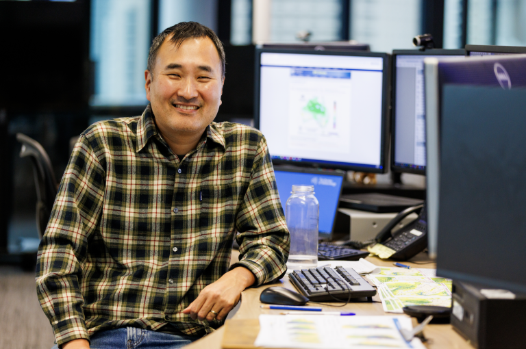 A man in a plaid shirt sits and smiles at a desk with multiple computer monitors and papers in an office setting.