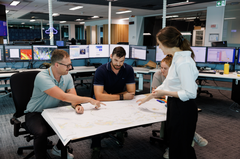 Four people in an office are discussing and pointing at a large map on a table, with computer monitors and office equipment in the background.
