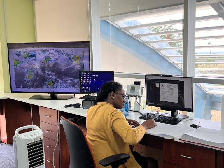 A person in a yellow sweater works at a desk with two monitors; one displays a weather map, the other shows a document. A large window and portable air conditioner are visible.