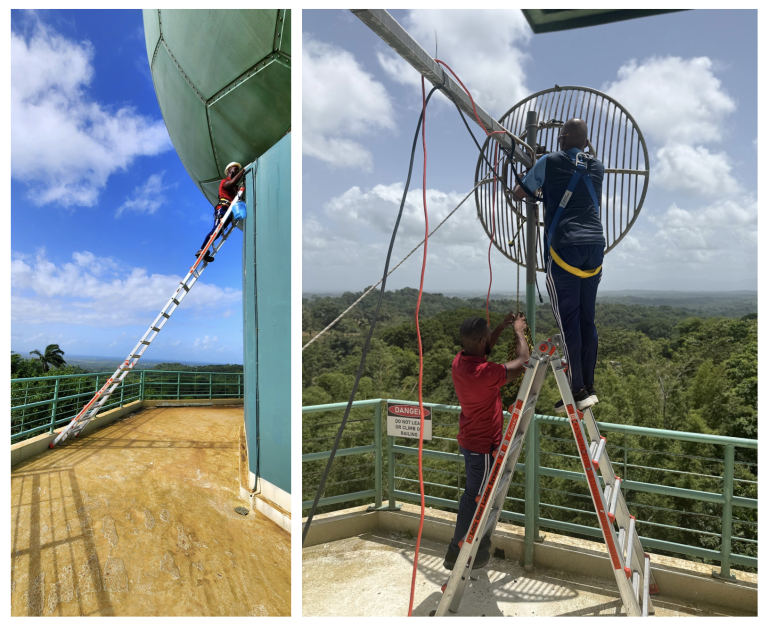 Two people on ladders install or adjust large equipment on a rooftop platform near a green tank, with cables and safety railings visible against a background of trees and sky.