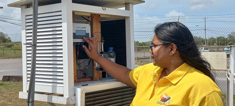 A woman in a yellow shirt adjusts a monitoring device inside a white outdoor enclosure near a fence and road under a partly cloudy sky.