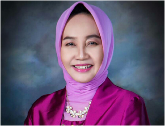 Woman wearing a pink hijab, pink outfit, and pearl necklace, smiling in front of a blue and gray studio background.