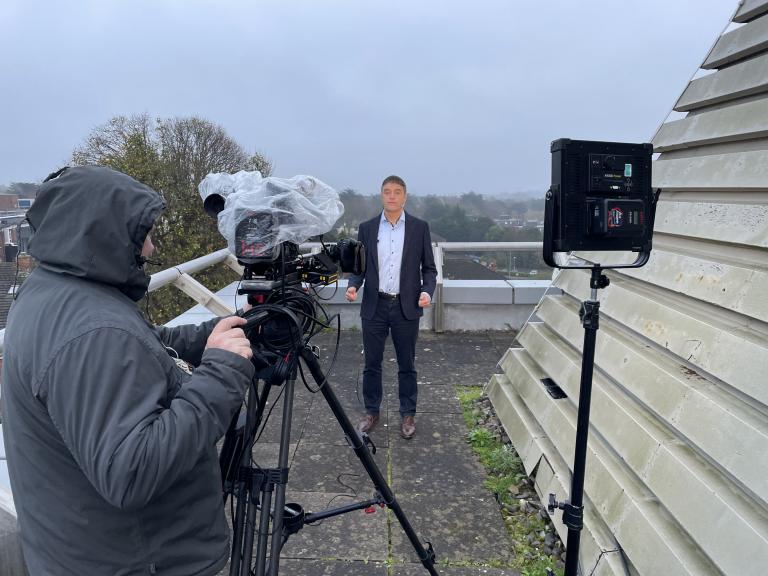 A man in a suit stands on a rooftop being filmed by a camera operator; professional lighting equipment is set up, and the weather appears overcast.