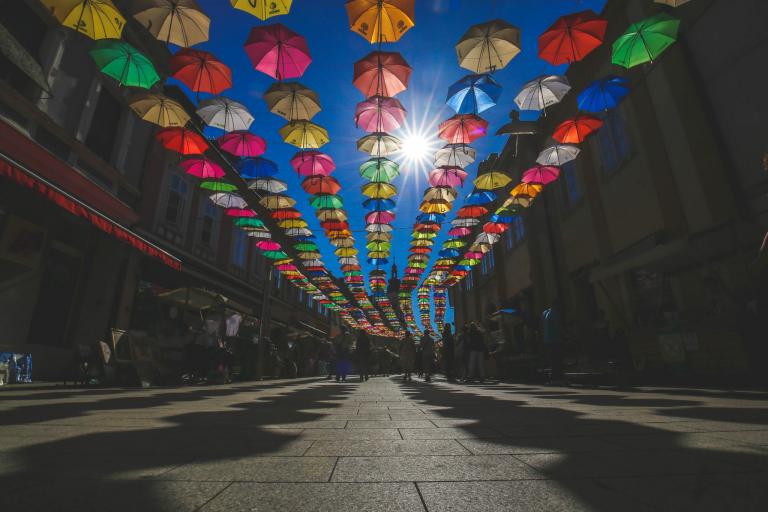 Rows of colorful umbrellas hang above a pedestrian street, casting shadows on the ground, with the sun shining brightly in a clear blue sky.