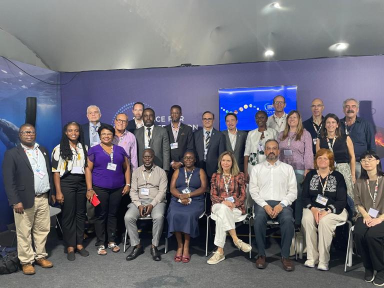 A group of conference attendees pose together indoors, some seated and others standing, all wearing name badges.