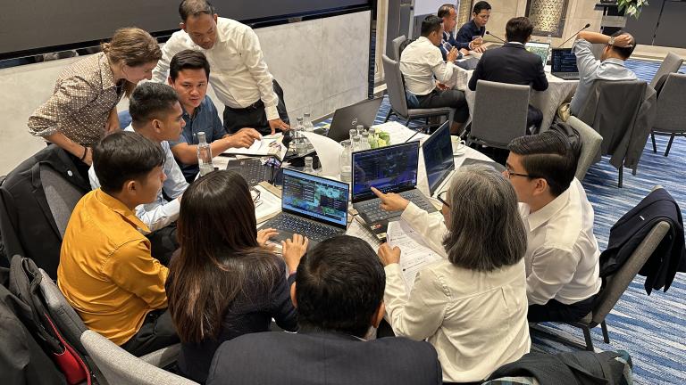 A group of people sit and stand around a table, collaborating and working on laptops during a business meeting or workshop.