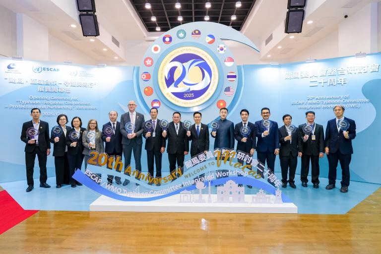 A group of people in formal attire stand in a row holding glass awards in front of a blue backdrop for the ESCAP/MMO 20th anniversary event.