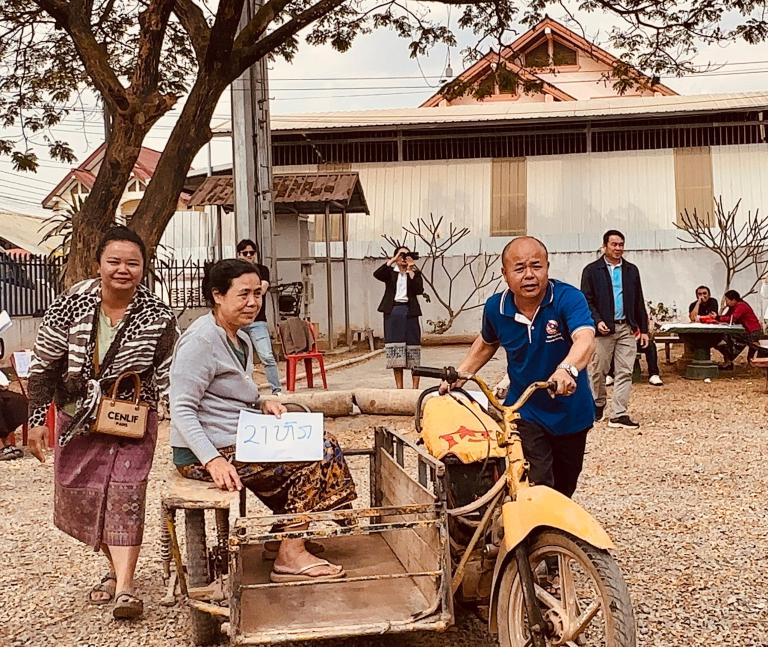A man pushes a woman seated on a wooden cart attached to a yellow tricycle; other people stand and watch in an outdoor setting. The woman holds a sign with Thai writing.