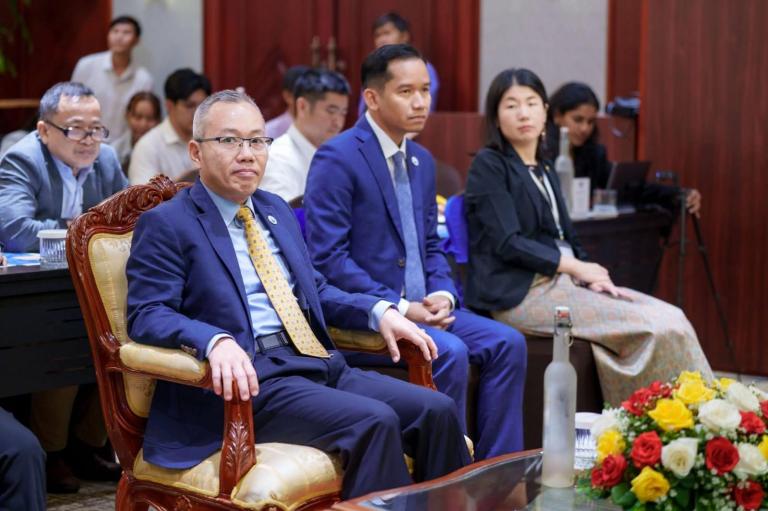 Several people in formal attire sit attentively in a conference room, with a bouquet of flowers and a water bottle on the table in the foreground.