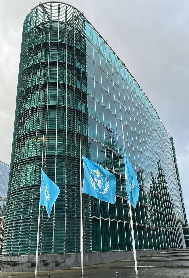 A modern glass building with three United Nations flags at half-mast in front, captured on a cloudy day.