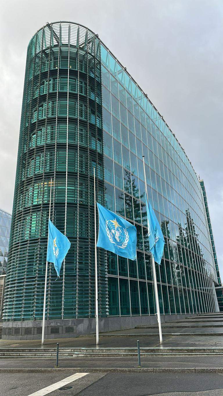 A modern glass office building with three United Nations flags flying at half-mast in front.