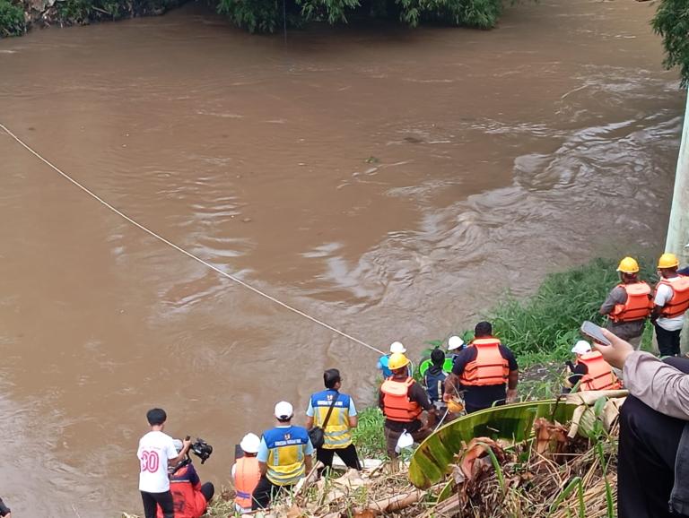 A group of rescue workers in life jackets and helmets stand by a muddy riverbank, some preparing equipment while others observe the water.
