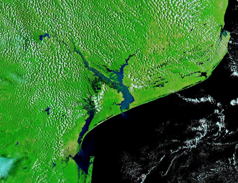 Satellite image showing a green coastal region with a river delta and estuary emptying into a dark blue ocean; scattered white clouds cover parts of the land and water.