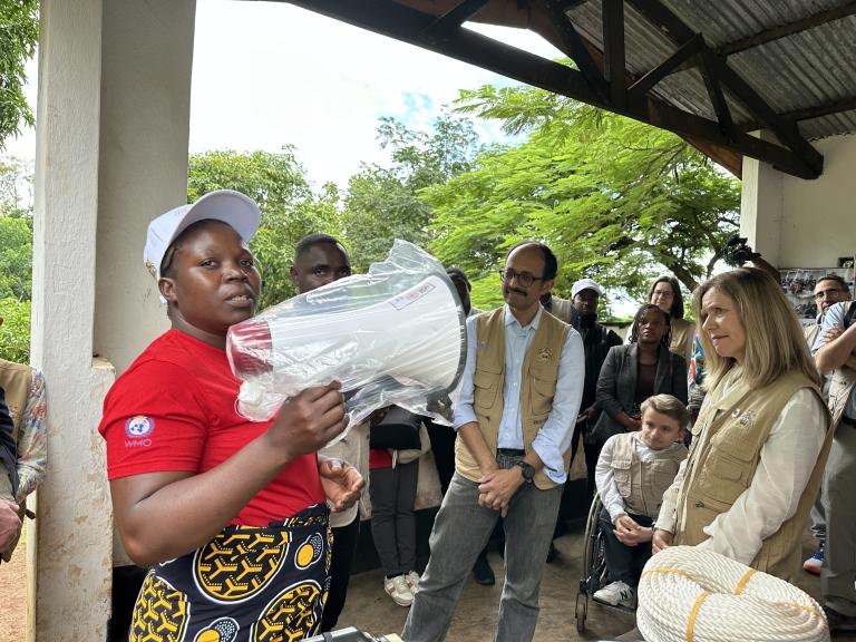 A woman in a red shirt holds a packaged medical device while speaking to a group of people standing and listening under a shelter.