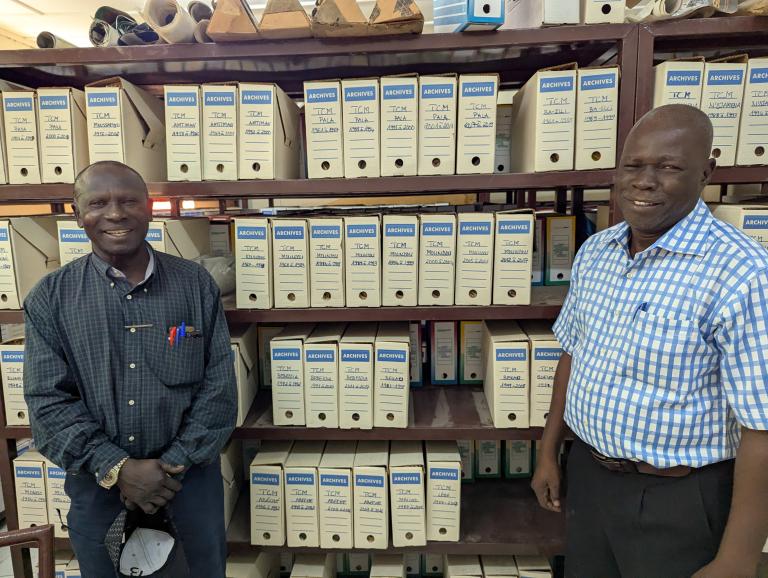 Two men stand in front of shelves filled with labeled archive boxes in an office or storage room.