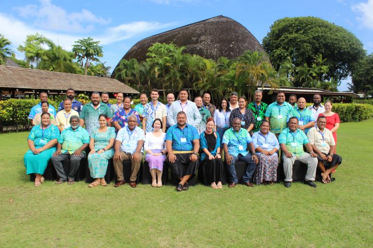 A large group of people pose for a photo outdoors in front of tropical greenery and a traditional wooden building under a blue sky.