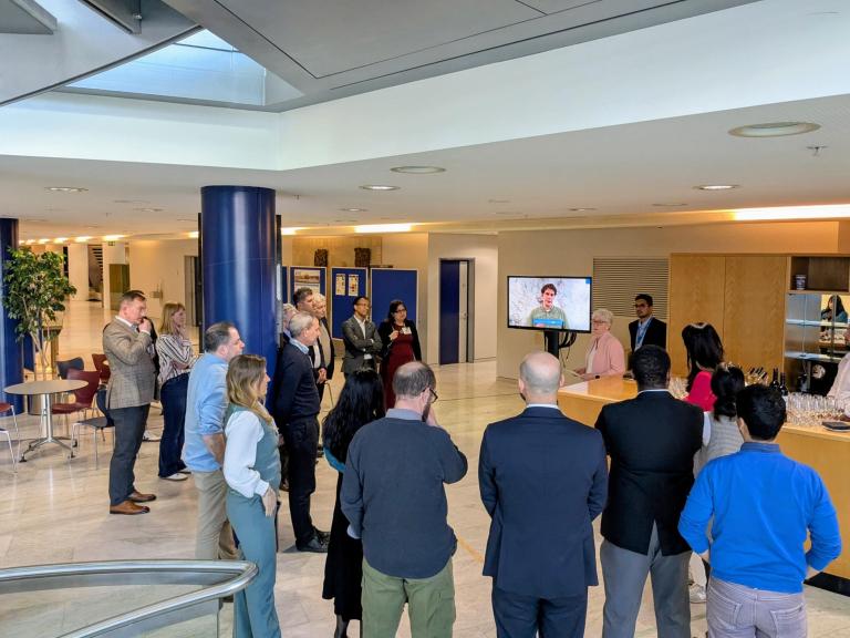 A group of people stands in an office lobby, watching a presentation on a TV screen where a person is speaking. Some refreshments are set up on a counter nearby.