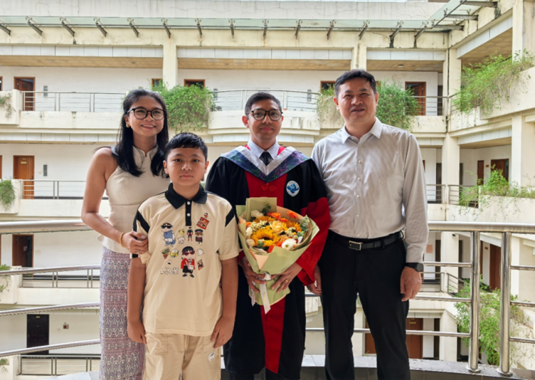 Four people pose together on a balcony; one person in the center wears a graduation gown and holds a bouquet of flowers, flanked by three others in casual and formal attire.