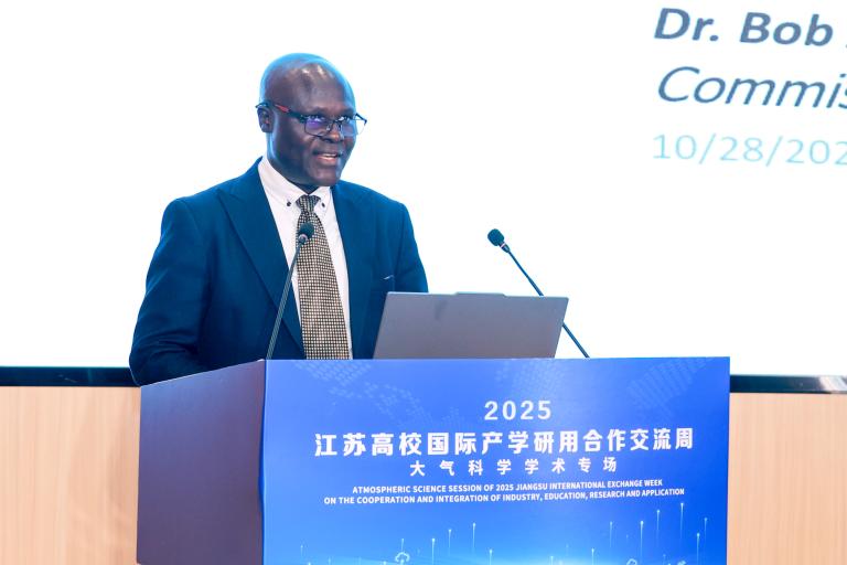 A man in a suit speaks at a podium during the 2025 Jiangsu International Exchange event, with a blue banner and presentation screen in the background.