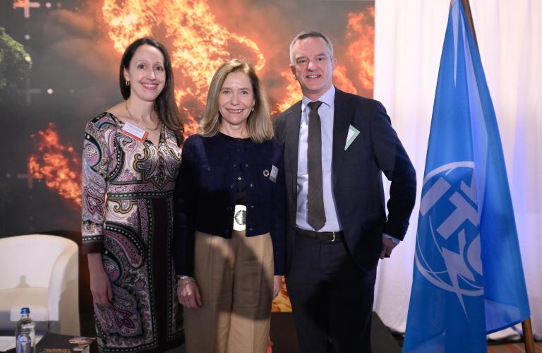 Three people pose together indoors beside a blue flag, with a fiery backdrop behind them and a table with a water bottle to the side.