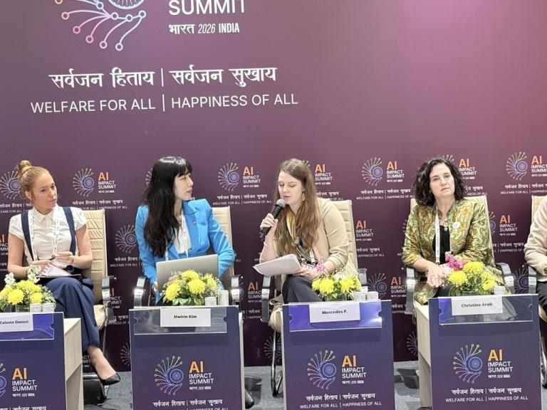 Four women sit on a panel at the AI Impact Summit 2024, with microphones and notes, against a backdrop displaying the event name and slogan.