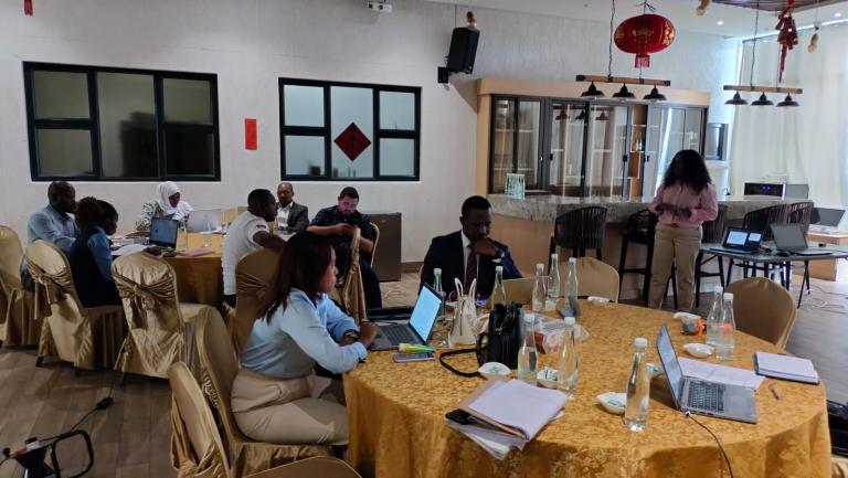 Several people sit at round tables with laptops and documents in a meeting room, while one person stands and presents near a screen.