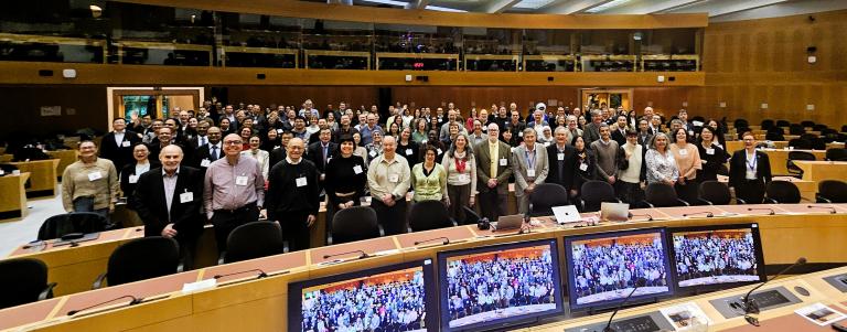 20260413_182748-2.jpg A large group of people pose for a photo in a conference room, with monitors in the foreground displaying the same scene.