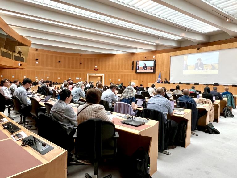 People seated in a large conference room attend a meeting, with speakers visible on a screen at the front and natural light coming in through the ceiling windows.