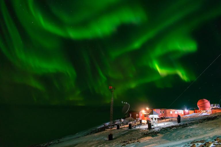 Green aurora borealis lights swirl in the night sky above a snowy research station with illuminated buildings and equipment.