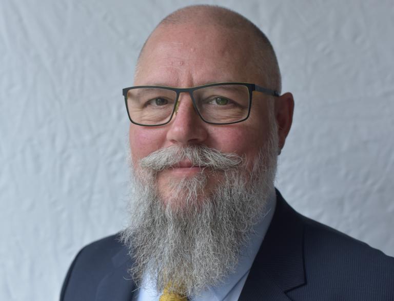 Bald man with glasses, a long grey beard, and a mustache, wearing a navy suit, light blue shirt, and yellow tie, against a plain light background.