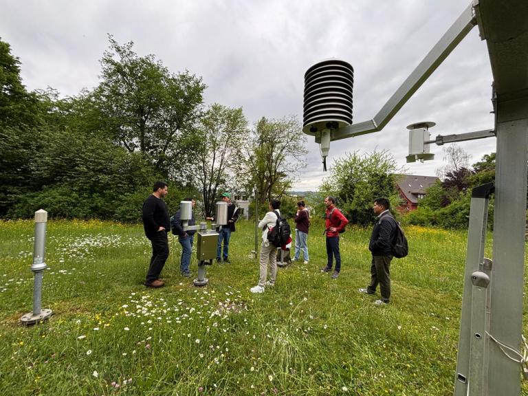 WhatsApp Image 2026-05-06 at 3.14.30 PM.jpeg A group of people stands in a grassy field examining weather instruments, including a Stevenson screen and rain gauges, surrounded by trees under a cloudy sky.