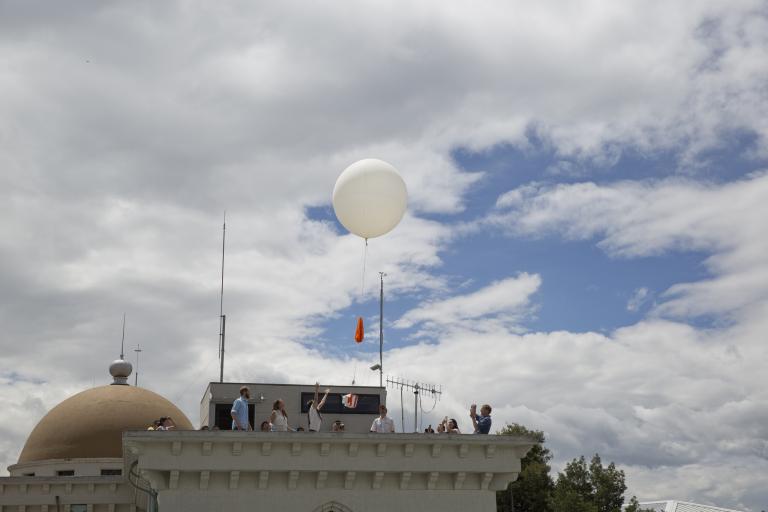 A group of people on a rooftop release a large white weather balloon into a partly cloudy sky.