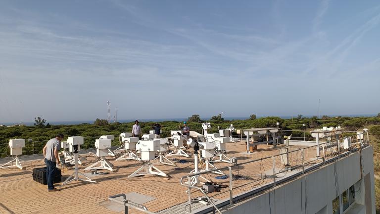 04. Ozone - Image 2.jpg Several people work among multiple white scientific instruments mounted on tripods on a rooftop, with trees and the sea visible in the background under a clear sky.