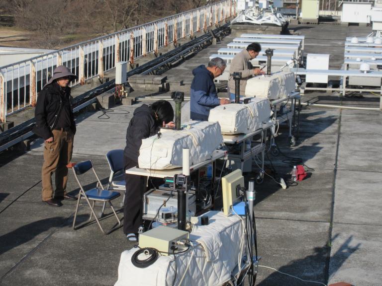 Several people work with covered scientific instruments on a rooftop, surrounded by technical equipment and chairs, under clear weather.