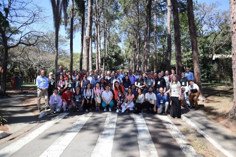 A large group of people poses for a photo outdoors on a crosswalk surrounded by tall trees and greenery.