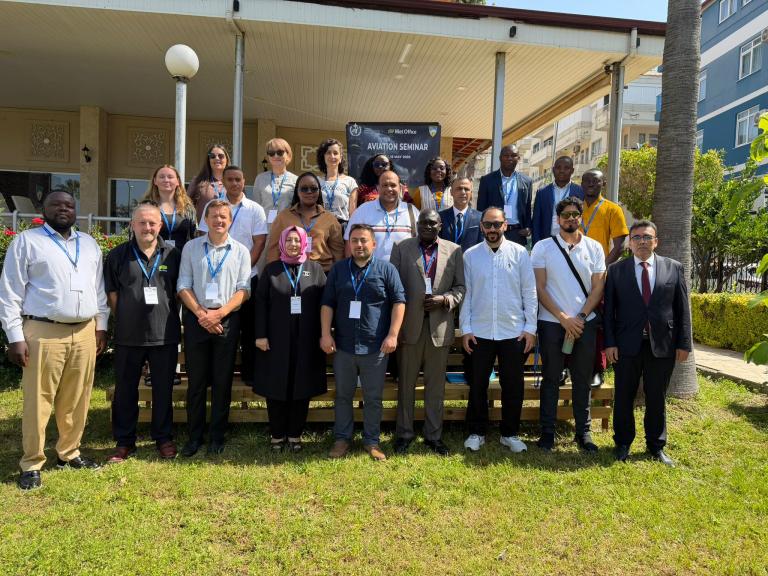 A group of people pose for a photo outdoors in front of a building, many wearing name badges, likely attending an aviation seminar.