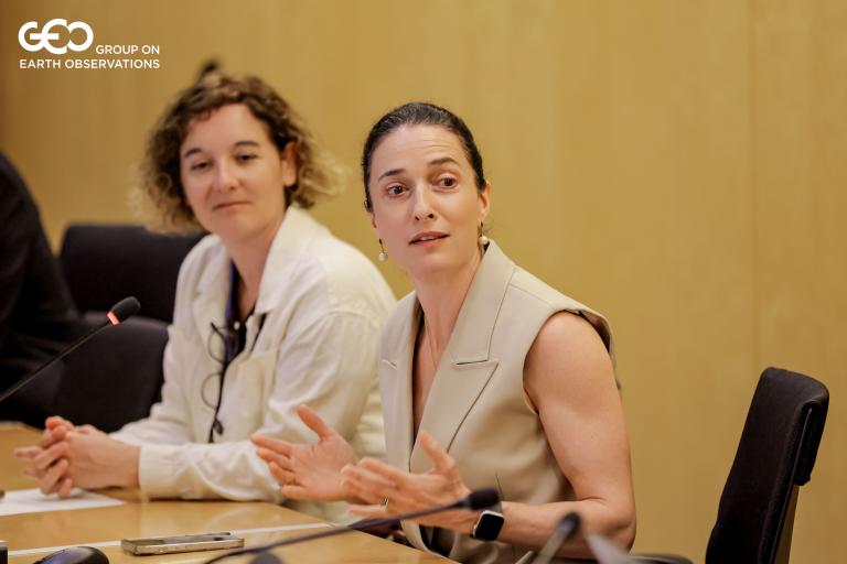 55311556819_664e4c57cb_o.jpg Two women sit at a conference table, one speaking while the other listens. Both have papers and a microphone in front of them. The background features a plain wooden wall.