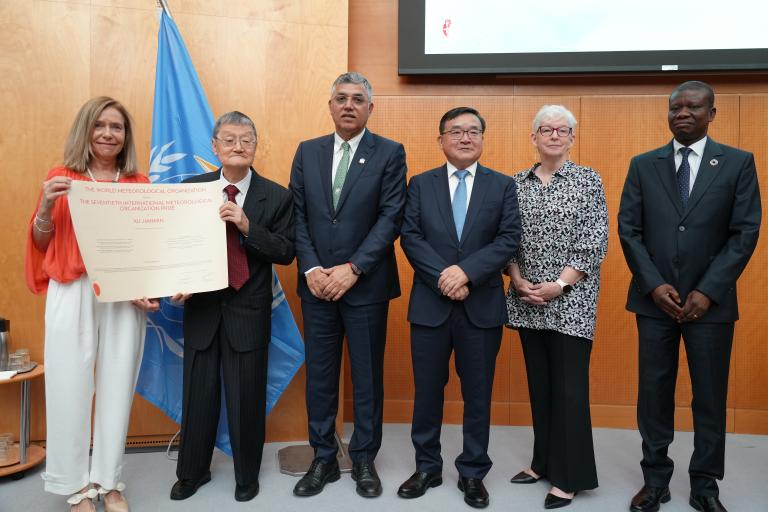 55353319832_3c023146b3_o.jpg Six people stand indoors; one person on the left holds a large certificate. A United Nations flag is visible in the background.