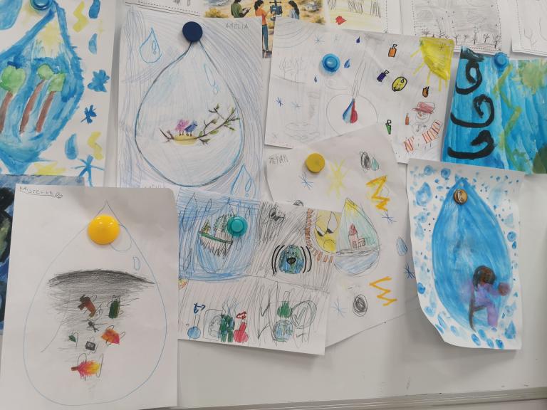 Children's drawings of water droplets, featuring various scenes and objects, are pinned to a whiteboard with colorful magnets.