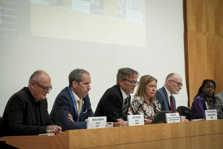 Six panelists sit at a long table during a conference, each with nameplates and microphones, against a plain backdrop with a projection screen above them.