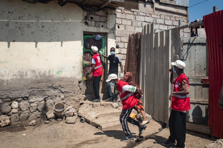 Red Cross workers in red vests walk toward a house with stone and brick walls; one person carries a child and another man stands by the door.