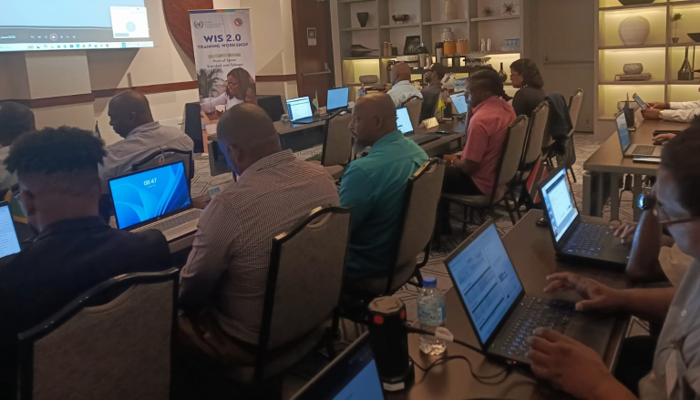 People seated in a classroom setting work on laptops during a training session; a presenter is at the front near a screen displaying a presentation.