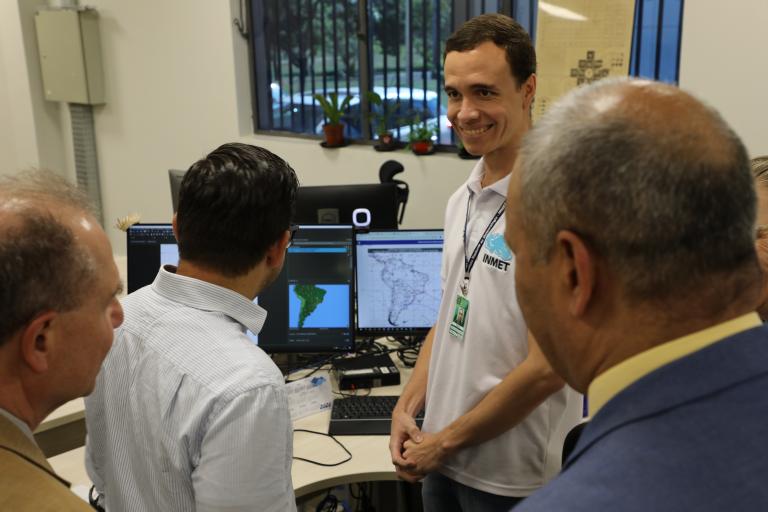 Four men stand around a workstation with computer screens displaying maps of South America. One man in a white shirt faces the camera and smiles.