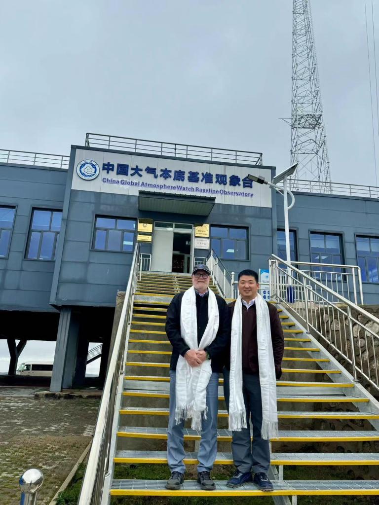 Two men in white scarves stand on stairs outside the China Global Atmosphere Watch Baseline Observatory building, with a tall antenna visible in the background.