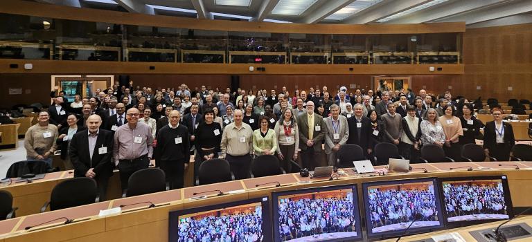 A large group of people poses for a photo inside a conference room, with several monitors in the foreground displaying the same group image.