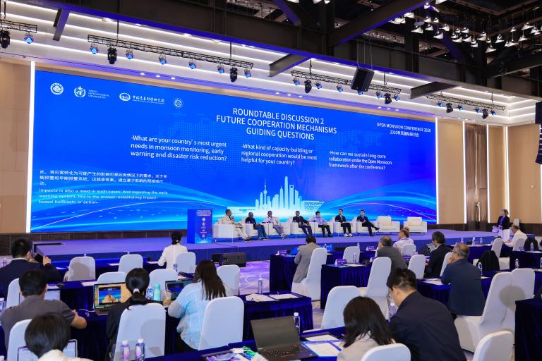 A conference room with a panel of speakers on stage, a large blue screen with guiding questions, and attendees seated at tables facing the stage.