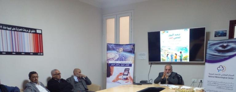 A group of men sit around a conference table in an office, with meteorological posters and a screen displaying a presentation in Arabic.