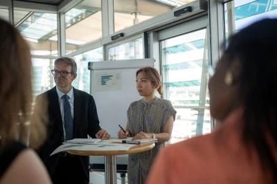 Two people stand at a table with papers and a pen, presenting near a flipchart with graphs, while others listen in a modern office setting.