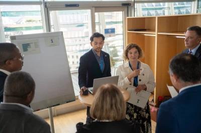 A group of professionals stand around a table with a laptop, discussing next to a flip chart in a modern office setting.